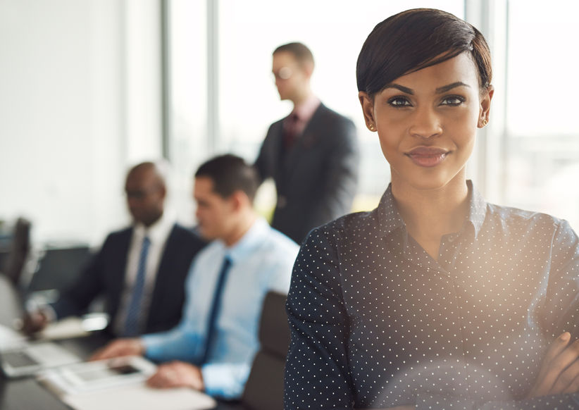 Young grinning business owner in office with polka dot blouse, folded arms and confident expression in front of group of mentees at conference table