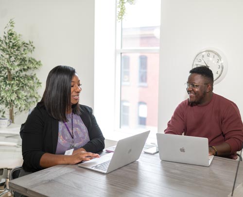 Two employees sitting in an office at computers discussing their mutual mentoring experience. 