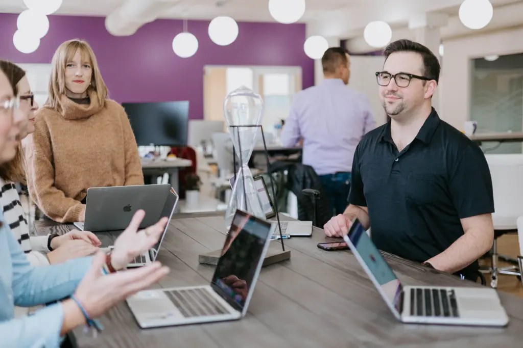 Employees sitting at a long table in an office with purple theme walls engaged in collaborative engagement discussions. 