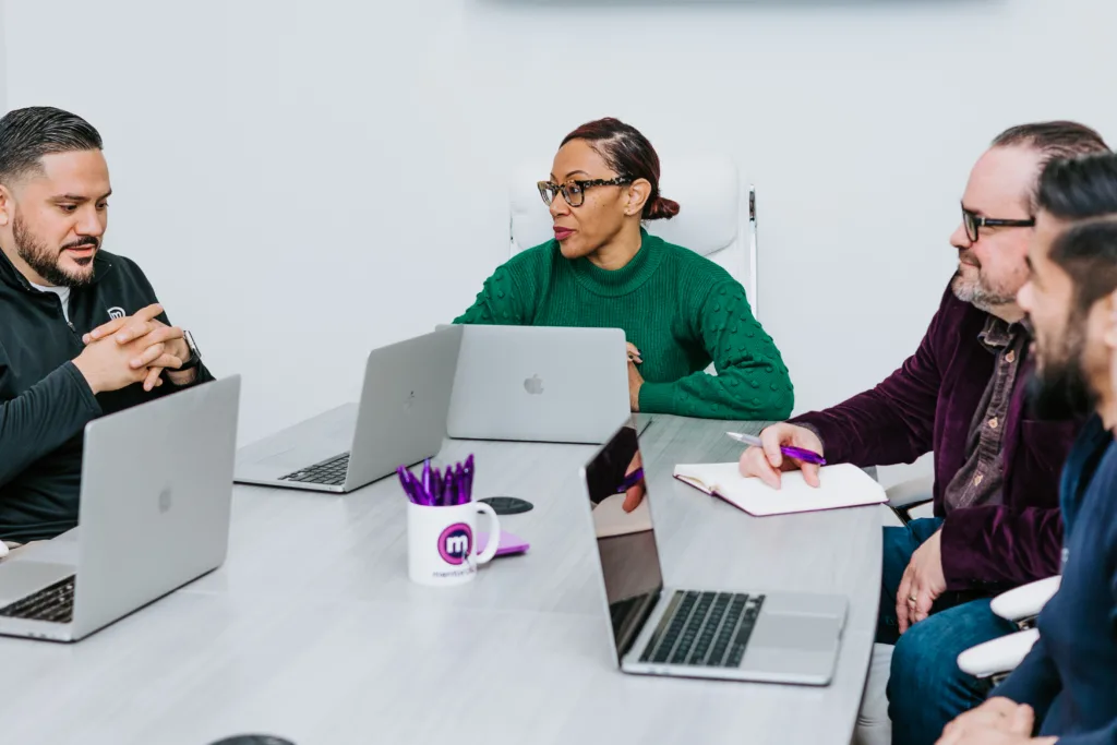 A team of workers sitting at a long gray table with computers talking about a succession plan. 